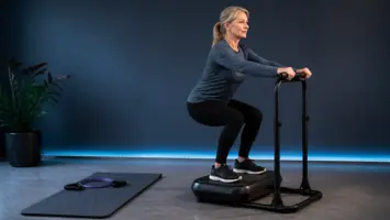 Kvinde laver squat på vibrationsplade med støttegelænder